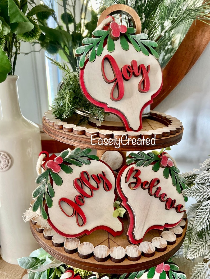 May include: Three wooden Christmas ornaments with red and green accents. The ornaments are shaped like teardrops and have the words "Joy", "Jolly", and "Believe" written on them in red.