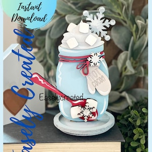 Könnte beinhalten: Ein Holzhandwerksprojekt, das ein blaues Einmachglas mit weißer Schlagsahne und Marshmallows, einen roten Löffel und einen Schneeflocke obenauf zeigt. Das Glas hat ein rotes Band und ein Etikett mit der Aufschrift "Hot Cocoa".