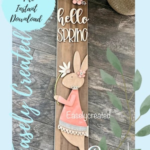 Puede incluir: Un archivo de corte digital para un letrero de madera con un conejito sosteniendo una flor y el texto "hello spring". El conejito lleva un vestido rosa y tiene una nariz y ojos marrones. El letrero es marrón y tiene una textura de madera.