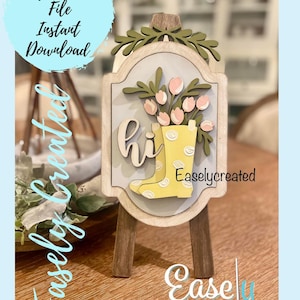 Puede incluir: Un letrero de madera con una bota de lluvia amarilla y flores rosas, con la palabra "hi" delante de la bota. El letrero está en un pequeño caballete de madera. El letrero dice "Easlycreated" en la parte inferior.