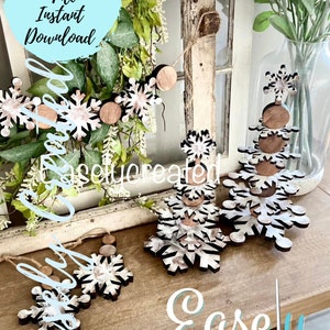 May include: A set of wooden snowflake ornaments with a distressed white paint finish. The ornaments are layered to create a tiered snowflake tree design. The ornaments are displayed on a wooden surface with a green plant in the background. The text "Digital Cut File Instant Download" and "Easily Created" are visible in the image.