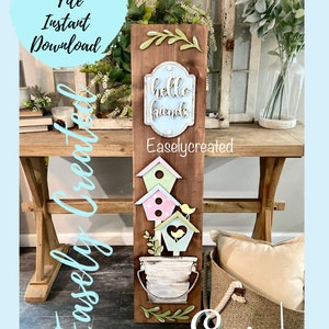 Puede incluir: Un letrero de madera con un letrero azul y blanco que dice "hello friends" y un diseño de tres casas para pájaros con un cubo de flores. El letrero está sobre una mesa de madera con una cesta de mimbre marrón y una almohada blanca.