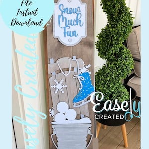 May include: Wooden winter-themed sign with the text "Snow Much Fun." The sign features a sled, ice skate, snowflakes, and a bucket of snowballs. The sign also has the text "Digital Cut File Instant Download."