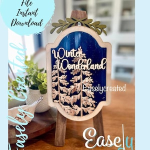 May include: A wooden sign with a blue background and gold glitter. The sign says "Winter Wonderland" and has a gold tree design. The sign is on a small wooden easel.