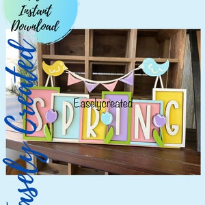 May include: A wooden sign spelling "SPRING" in colorful letters with a yellow and blue bird on each end of a string of flags. The sign is on a wooden shelf.