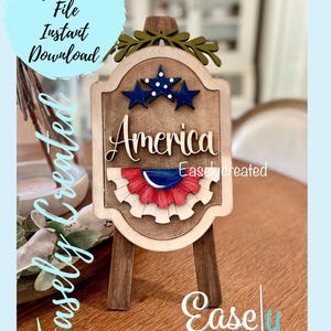 May include: A wooden sign with the word "America" on it, decorated with stars and a red, white, and blue ribbon. The sign is on a small easel.