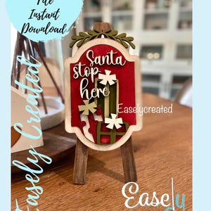 May include: A wooden sign with a red background and the text "Santa stop here" in white. The sign is decorated with green leaves and snowflakes. The sign is on a small wooden easel.