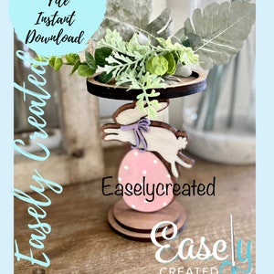 May include: Wooden Easter-themed decoration featuring a bunny jumping over a pink polka-dotted egg. The decoration has a tiered design with greenery and the text "Easely Created" and "Digital Cut File Instant Download".