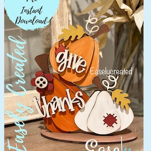 May include: A wooden craft project featuring three pumpkins with the words "give thanks" on them. The pumpkins are decorated with fall leaves and a red flower. The project is on a wooden base.