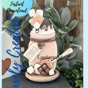 Może przedstawiać: Drewniany projekt rzemieślniczy słoika wypełnionego wzorem w stylu s'mores. Słoik jest ozdobiony czekoladą, piankami i drewnianą łyżką. Słoik ma etykietę z napisem "S'mores" a na słoiku nadrukowane są słowa "Easlycreated".
