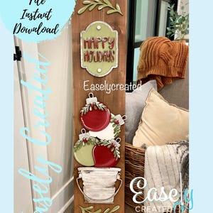 May include: A wooden sign with a green and white "Happy Holidays" sign, three red and green Christmas baubles stacked in a white bucket, and green leaves on a brown background. The sign is labelled "Easliycreated".