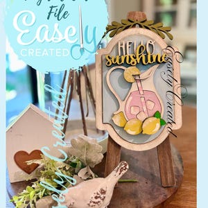 May include: A wooden sign with the text "Hello Sunshine" and a pitcher of pink lemonade with lemons. The sign is on a small wooden easel. Other decorative items include a small house, a bird figurine, and greenery. The words "Easely Created" are also visible.