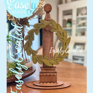 May include: A wooden decorative piece featuring a central pillar with a circular olive branch wreath. The piece is brown with a light green wreath and a round base. Text on the image reads "Digital Cut File Easely Created".