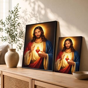 Two sizes of the Sacred Heart of Jesus canvas prints displayed side-by-side on a wooden credenza. The beautiful Christian artwork showcases rich red and blue robes with glowing golden light textures. Perfect size guide for customers looking to choose religious wall art for their home altar, bedroom, or parish.