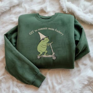 May include: Green sweatshirt with the phrase "Off to commit mom foolery" above a cartoon frog riding a scooter. The frog wears a wizard hat. The sweatshirt is a solid, dark green color.