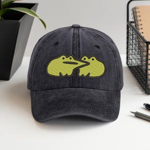 Pode incluir: Boné de beisebol preto com acabamento desgastado. O boné apresenta um design bordado de duas rãs verde claro voltadas uma para a outra. As rãs têm contornos pretos e estão posicionadas na frente do boné.