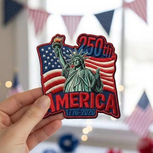 May include: Embroidered patch featuring the Statue of Liberty, the American flag, and the text "250th AMERICA 1776-2026". The patch has a red, white, blue, and green color scheme. The patch is held by a hand.
