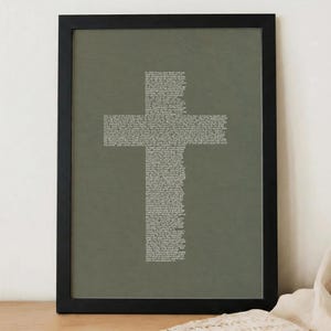 May include: A framed wall art piece featuring a dark olive green background with white text arranged in the shape of a cross. The text consists of numerous lines of small, dense script that fill the entire cross silhouette within the black frame.