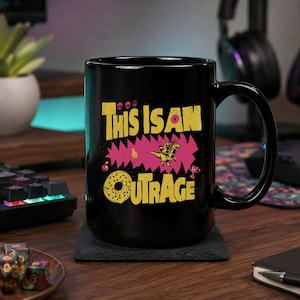 May include: A black ceramic mug featuring a yellow graphic design with the text "THIS IS AN OUTRAGE". The design includes a small dinosaur, a donut, three skulls, a pineapple, a bomb, and a trophy, all set against a jagged pink background.