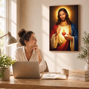Sacred Heart of Jesus framed canvas print hanging in a bright home office. A woman sits at a wooden desk with a laptop, looking thoughtfully at the beautiful Catholic artwork. On the desk are spiritual books, a mug, and a small green plant. Perfect Christian office decor, Catholic gift for women, or inspirational daily study room art.