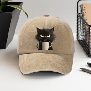 May include: Tan baseball cap featuring a cartoon black cat with an angry expression, holding a white coffee mug. The cat is the central design element, set against the neutral background of the cap. The cap has a curved brim and a button on top.