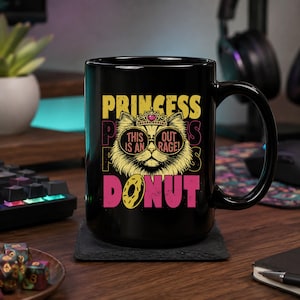 Puede incluir: Una taza de cerámica negra con el gráfico de un gato que lleva una corona y gafas de sol. El texto dice PRINCESS en la parte superior, THIS IS AN OUT RAGE! en las gafas y DONUT en la parte inferior. El gato es amarillo con un corazón rosa.