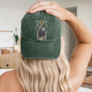 A woman wearing a green washed cotton cap featuring an intricate embroidery of Christ seated on a throne. The cap symbolizes faith, offering a stylish and spiritual accessory for casual outings. With an adjustable strap, this hat ensures a comfortable fit and is perfect for expressing your Christian beliefs or gifting to loved ones. Ideal for church, daily wear, or as a meaningful token for those who embrace their faith.