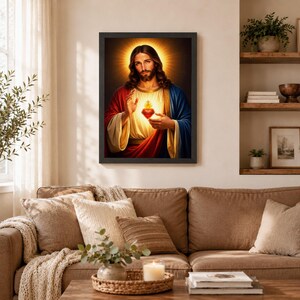 Sacred Heart of Jesus framed portrait in a black frame, hanging above a comfortable brown sofa with cozy throw pillows. The warm living room setting includes wooden shelves with books and small plants. Elegant Catholic wall decor that brings divine peace, protection, and blessing to your family's main living space.