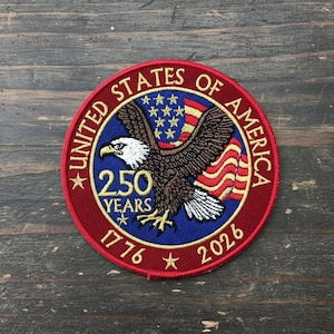 May include: A round embroidered patch with a patriotic theme. The patch features a bald eagle with outstretched wings, the American flag, and the text "UNITED STATES OF AMERICA" and "250 YEARS 1776-2026". The patch is red, white, blue, and brown.