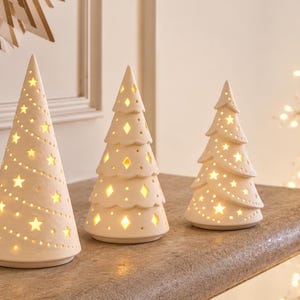 May include: Three off-white ceramic tree-shaped luminaries of varying heights. The tallest cone-shaped tree features star-shaped cutouts, while the two shorter, tiered trees feature diamond and star-shaped cutouts, all emitting a warm internal glow.