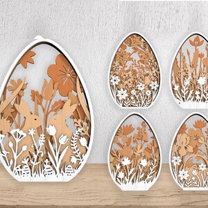 May include: Wooden Easter egg decorations with intricate laser-cut designs. The eggs feature floral and bunny motifs in shades of brown and white. The designs include flowers, bunnies, and foliage, creating a layered, three-dimensional effect. These decorative eggs are perfect for Easter.