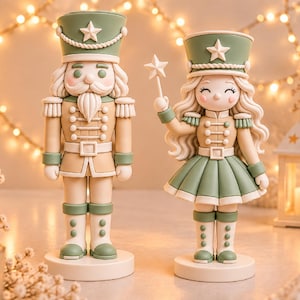Puede incluir: Dos figuras de cascanueces en tonos verde salvia y crema. La figura de la izquierda es un soldado con bigote y uniforme. La figura de la derecha tiene el pelo largo y ondulado y sostiene una varita con una estrella. Ambas sobre bases.