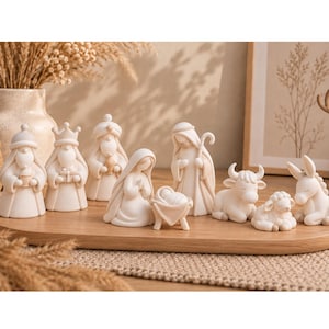 Puede incluir: Un conjunto de belén con figuras de color crema de la sagrada familia, los tres reyes magos y animales, incluyendo una vaca, una oveja y un burro. Las figuras son lisas, estilizadas y están dispuestas sobre una bandeja de madera ovalada.