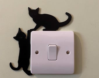 May include: A white light switch with a rectangular design, featuring two black cat silhouettes. One cat is perched above the switch, while the other appears to be climbing. The cats add a decorative touch to the functional switch.