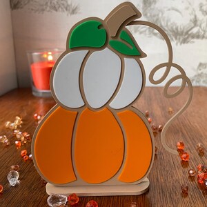 May include: A layered pumpkin decoration featuring a tan base, a white upper section, and an orange lower section. The design includes two green leaves at the top, a tan stem, and a tan curly vine extending to the right side. The object stands upright.