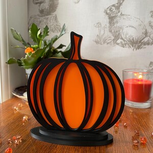 May include: A layered pumpkin decoration featuring alternating black and bright orange panels. The object has a central stem and is mounted on a solid black oval base, creating a dimensional effect with its stacked, cutout design.