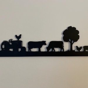 May include: Black metal farm scene silhouette depicting a tractor, cows, a tree, a chicken, and a pig. The artwork is mounted on a white wall, creating a contrast between the dark silhouette and the light background. The design evokes a rural, country theme.