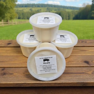 Peut inclure: Quatre pots en plastique blanc de saindoux, empilés sur une surface en bois. Chaque pot est muni d'un couvercle blanc et d'une étiquette avec le texte "PIG LARD" et une illustration de porc. En arrière-plan, un champ vert et des arbres.