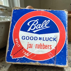 May include: A vintage box of Ball jar rubbers. The box is blue with a red and white circular design. The Ball logo is in white script. The words "GOOD LUCK" and "jar rubbers" are also visible. The box has a split tab.
