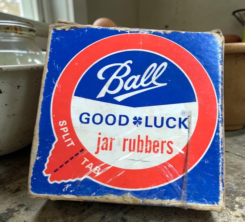 May include: A vintage box of Ball jar rubbers. The box is blue with a red and white circular design. The Ball logo is in white script. The words "GOOD LUCK" and "jar rubbers" are also visible. The box has a split tab.