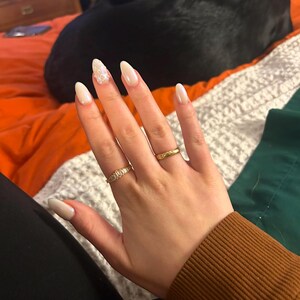 May include: A hand with white almond-shaped nails with glitter on one nail. The hand is wearing two gold rings.