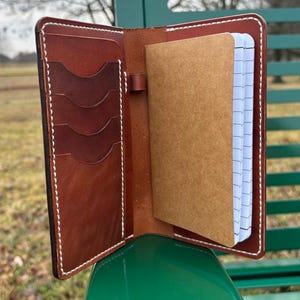 May include: Open brown leather notebook cover with white stitching. The cover has multiple card slots on the left side and a notepad on the right. The notepad has a brown cover and white lined pages.