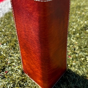 May include: A tall, rectangular leather item in a deep reddish-brown colour. The edges are stitched with white thread, providing a contrasting border. It is positioned on a green surface.