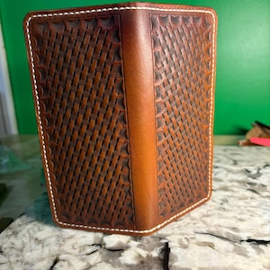 May include: A brown leather wallet with a woven basket pattern and white stitching. The wallet is rectangular and appears to be closed, with a vertical strip of leather on the spine. The background is a green wall and a marble countertop.