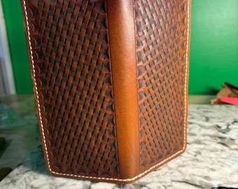 May include: A brown leather wallet with a woven basket pattern and white stitching. The wallet is rectangular and appears to be closed, with a vertical strip of leather on the spine. The background is a green wall and a marble countertop.