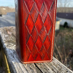 Puede incluir: Una funda de diario o cuaderno de cuero vertical, hecha a mano. La funda es de un rico color marrón rojizo con un patrón de diamantes y costuras blancas en los bordes. Está colocada sobre una superficie de madera.