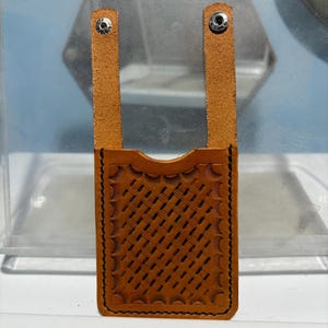 Könnte beinhalten: Ein hellbrauner Lederkartenhalter mit einem gewebten Muster und zwei Riemen, die mit Nieten befestigt sind. Die Kanten sind mit schwarzem Faden genäht. Der Kartenhalter ist für Karten und Bargeld konzipiert.