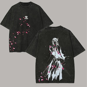 Könnte beinhalten: Dunkelgraues T-Shirt mit Grafikdesign. Auf der Vorderseite befindet sich ein kleines Totenkopf-Logo mit dem Wort "BNEAH" und rosa Blütenblättern. Auf der Rückseite ist eine weiß-blaue Illustration eines Charakters mit rosa Blütenblättern zu sehen.
