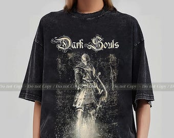 May include: Dark gray t-shirt featuring a distressed graphic of a knight with a sword and shield. The text "Dark Souls" is arched above the illustration in a decorative font. The shirt has a vintage, worn look.