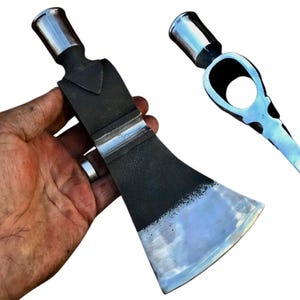 May include: A hand holds a black and silver tomahawk with a polished blade and a rounded head. The tomahawk has a hole in the head for a handle.